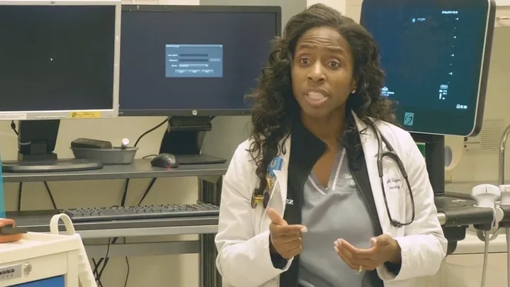 The image shows a healthcare professional, likely a doctor, standing in a medical environment. She is wearing a white coat over gray scrubs, with a stethoscope around her neck. The doctor is speaking and gesturing with her hands, possibly explaining something. Behind her are medical monitors and equipment, suggesting that she is in a clinical or hospital setting. The room has a technical atmosphere, with computers and diagnostic screens visible in the background.