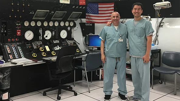 Two medical professionals in scrubs standing in a control room.