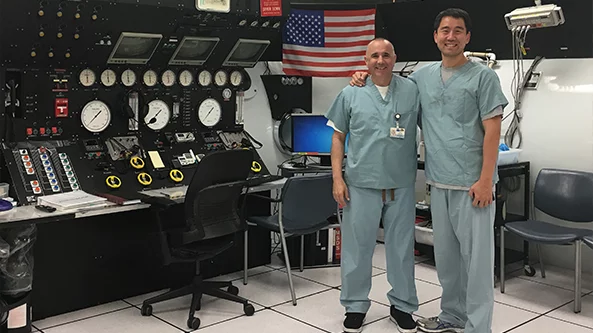 Two medical professionals in scrubs standing in a control room.
