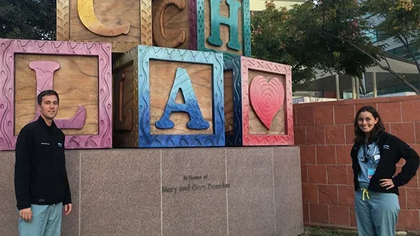 Two medical professionals standing in front of a colorful sculpture made of large blocks, each with a letter or symbol. The blocks spell out "CHLA" with a heart symbol, indicating the Children's Hospital Los Angeles.