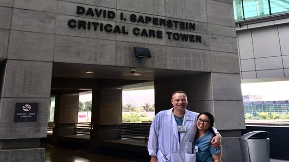 Two medical professionals standing in front of the entrance to the "David I. Saperstein Critical Care Tower" at Cedars-Sinai