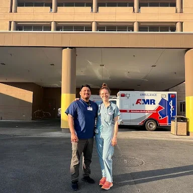 Two medical professionals stand smiling in front of a hospital entrance, with an AMR ambulance parked behind them. The person on the left is wearing a dark blue scrub top, while the person on the right is in light blue scrubs with orange shoes. The setting appears to be outside a hospital facility on a sunny day.