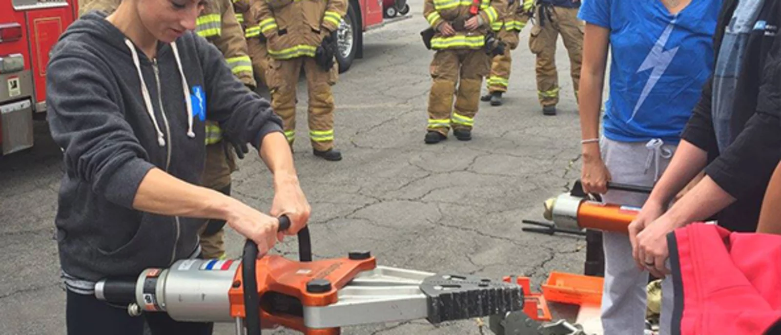 Group of people participating in a demonstration involving fire rescue equipment. A woman in the foreground is handling a large hydraulic rescue tool, commonly known as the "Jaws of Life".