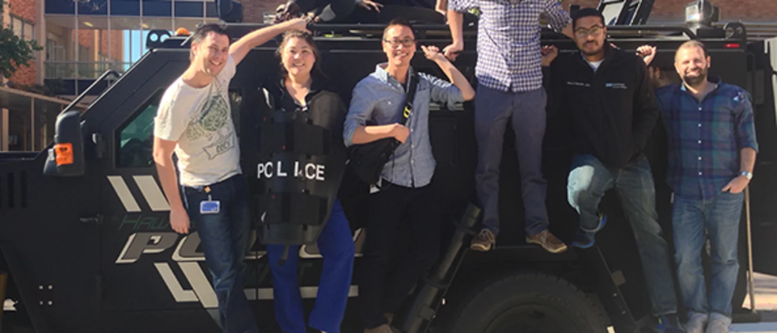 A group of eight people posing together on and around a black police SWAT vehicle.