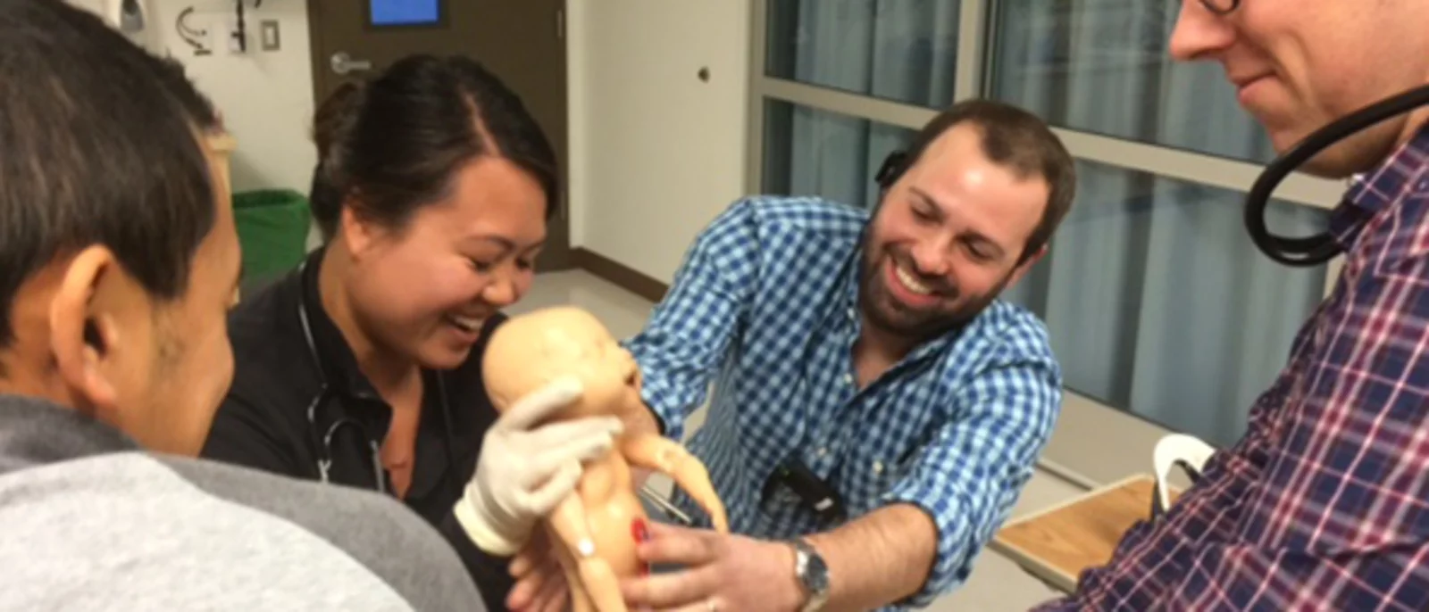 A group of medical professionals gathered around a mannequin, practicing a medical procedure. One person is holding a baby mannequin, while others are assisting and observing. The scene appears to be a training session, possibly in a hospital or clinical setting, with everyone focused and engaged.