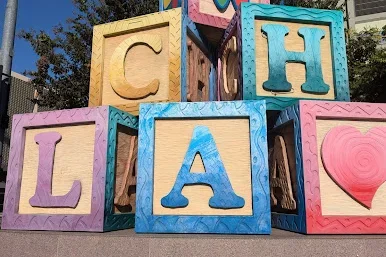 Colorful building block sculptures spelling "CHLA" stand outside the Children's Hospital Los Angeles, with decorative symbols like a butterfly and heart on some blocks. The hospital building is visible in the background.