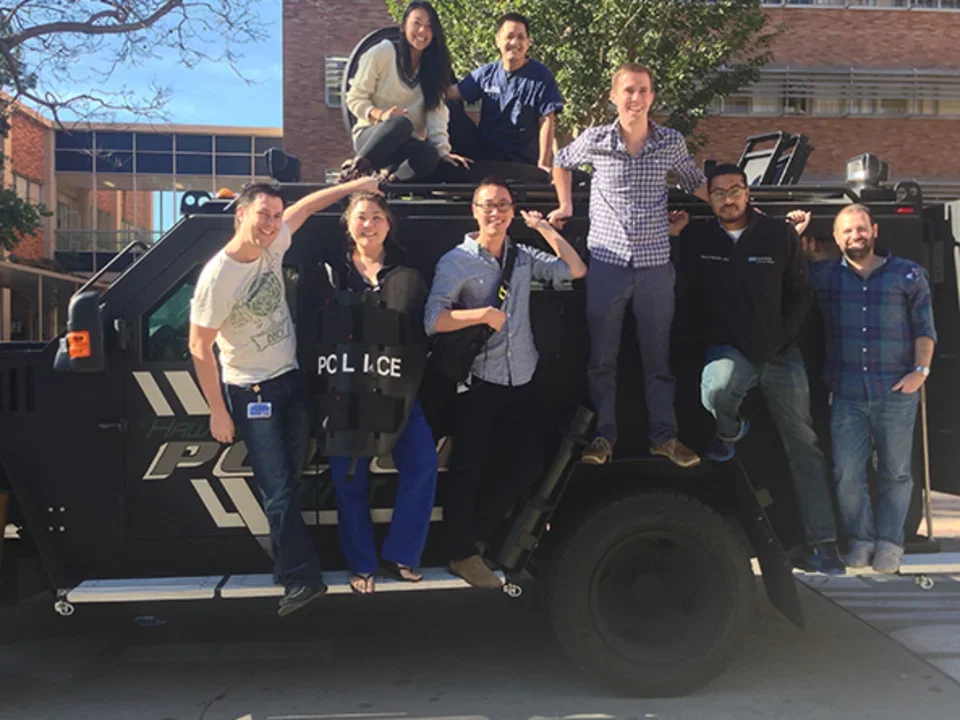 A group of eight people posing together on and around a black police SWAT vehicle.