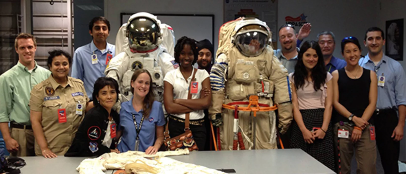 Group of people posing in a classroom or lab setting. Two individuals are wearing astronaut suits, while the others are dressed in casual or professional attire.