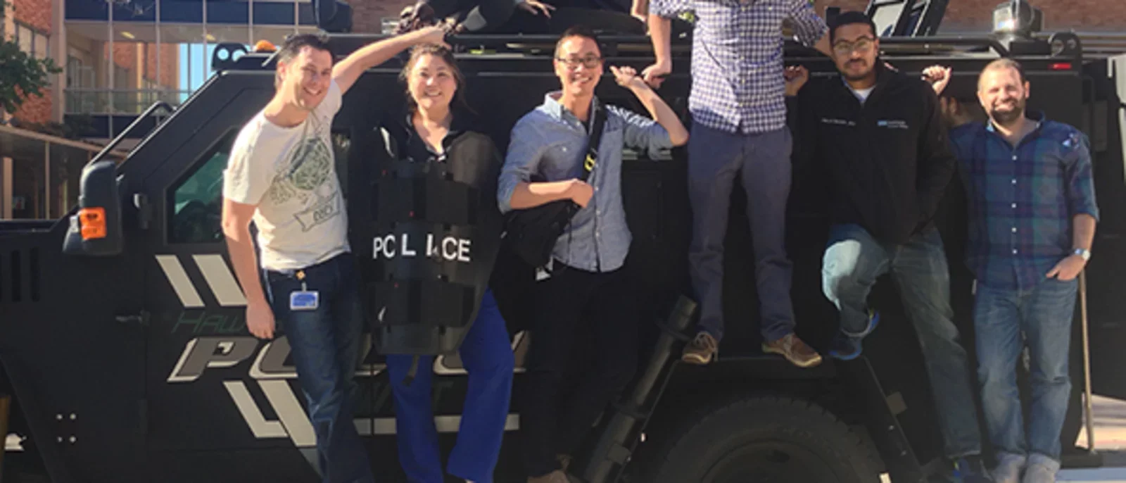 A group of eight people posing together on and around a black police SWAT vehicle.