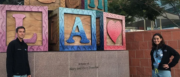 Two medical professionals standing in front of a colorful sculpture made of large blocks, each with a letter or symbol. The blocks spell out "CHLA" with a heart symbol, indicating the Children's Hospital Los Angeles.